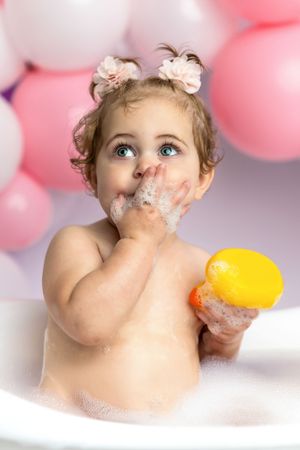 Cute baby girl in bubble bath holding yellow rubber duck with pink balloons in background.