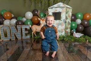 Happy toddler in denim overalls beside deer decor and ONE sign with woodland balloon backdrop.