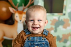 Smiling baby boy in denim overalls and brown shirt at a woodland themed birthday party with deer decorations.