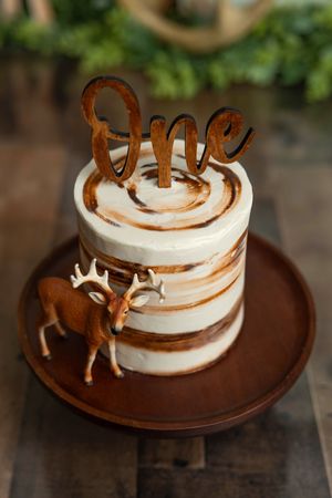 Rustic first birthday smash cake with deer figurine, wood "One" topper, and swirled frosting on brown plate.