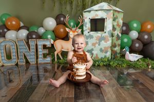 Baby boy smashes first birthday cake at hunting-themed party with deer, duck decoys, camo fort, and ONE sign.