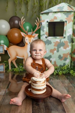 Baby boy in brown overalls smashes first birthday cake at woodland hunting themed party with deer and camo decorations.