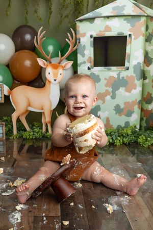 Baby smashing first birthday cake in camo-themed party setup with deer cutout and balloon decorations.