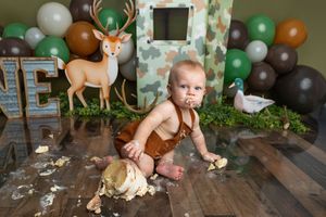 Baby boy in brown suspenders smashing his first birthday cake on a wood floor with deer and balloon decorations.