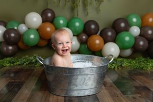Smiling baby sitting in a galvanized tub with milk, surrounded by green, brown, and white balloons on a wooden floor.