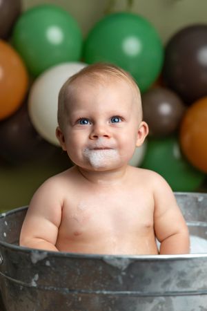Baby boy sitting in a tin tub with frosting on his face, surrounded by green, brown, and white balloons.