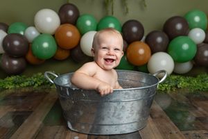 Laughing baby sitting in a vintage metal tub with green, brown, and white balloon backdrop.