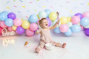 Baby girl joyfully celebrates her first birthday smash cake session with colorful balloons and a neon One sign.