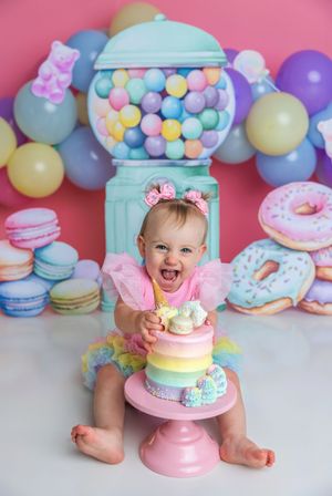 Baby girl in pink tutu smashes rainbow cake surrounded by colorful balloons, macarons, donuts, and a gumball machine backdrop.
