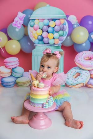Baby girl in rainbow tutu eating first birthday cake smash surrounded by colorful candy decorations and balloons.