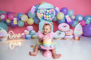 Baby girl smashing a pastel rainbow cake surrounded by donuts, macarons, balloons, and a gumball machine at a candy-themed first birthday.