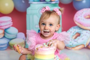 Baby girl in pink tutu smashing rainbow birthday cake, surrounded by colorful donut and macaron decorations.