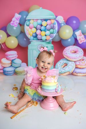 Laughing baby girl in pink ruffle tutu holds rainbow cake at vibrant candy-themed first birthday smash session.