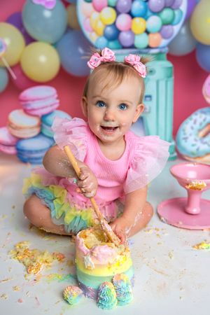 Baby girl in pink tutu smashes rainbow cake with spoon at colorful candy-themed first birthday photo shoot.