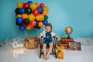 Baby in overalls sits among colorful balloons, globe, and suitcase in an adventure-themed first birthday photo.