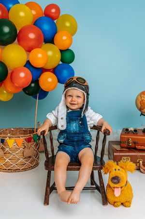 Smiling toddler in denim overalls and pilot hat sits on wooden chair with colorful balloons and stuffed dog toy.
