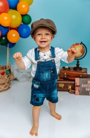 Smiling toddler boy in denim overalls and newsboy cap stands barefoot with colorful balloons and vintage suitcases behind him.