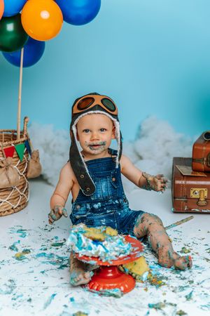 Baby in aviator hat smashes first birthday cake surrounded by balloons and vintage suitcases on blue background.