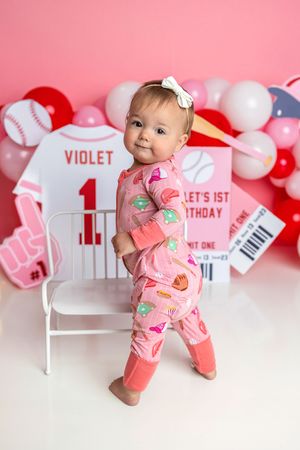 Baby girl Violet in pink pajamas stands at her baseball-themed 1st birthday photo shoot with pink balloons.