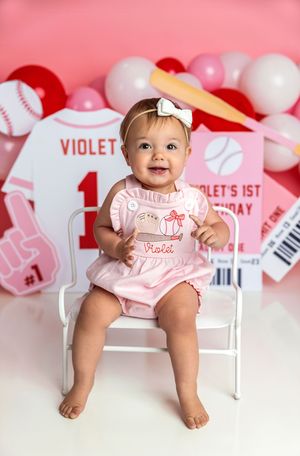 Baby girl Violet smiles in pink outfit at baseball-themed 1st birthday photo shoot with balloons and decorations.