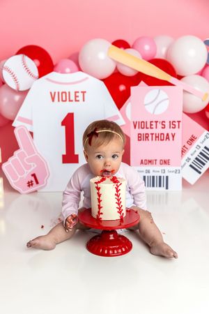 Baby girl enjoys her baseball-themed 1st birthday cake smash with pink and red decorations and personalized Violet jersey backdrop.