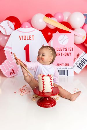 Baby Violet celebrates her 1st birthday smashing a baseball-themed cake, surrounded by pink balloons and sports decorations.