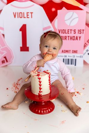 Baby Violet enjoys her baseball-themed smash cake during her 1st birthday celebration, surrounded by pink and red decorations.