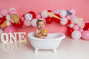 Baby girl sits in a white mini bathtub holding a rubber duck, surrounded by balloons and a lit ONE sign for her first birthday.