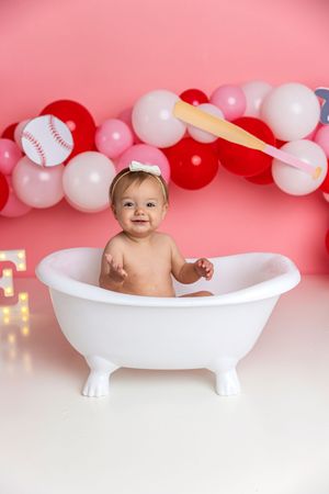 Smiling baby girl in white clawfoot tub with pink and red baseball balloon garland backdrop.