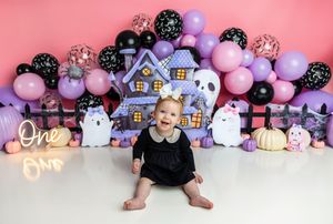 Baby girl in black dress sits before Halloween backdrop with purple house, balloons, and ghost decorations at first birthday cake smash.