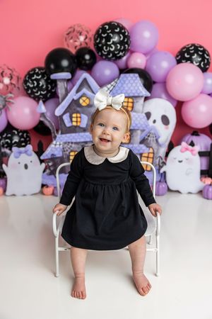 Baby girl in black dress with white bow sits before Halloween balloon backdrop with ghosts and haunted house decorations.