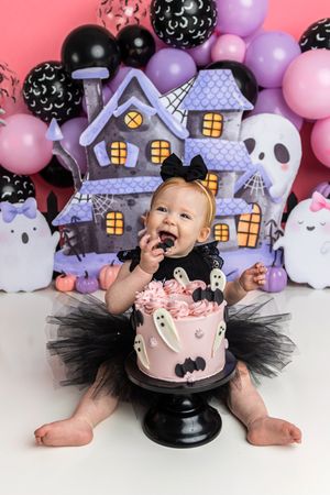 Baby girl in black tutu smashes Halloween-themed first birthday cake with ghost and bat decorations amid spooky balloon backdrop.