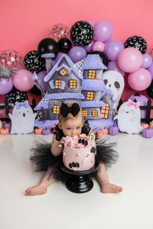 Baby girl in black tutu smashes pink Halloween cake surrounded by ghost decorations and purple balloons.