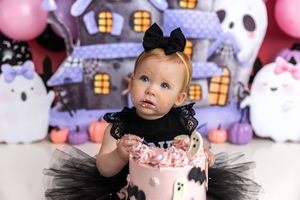 Baby girl in black tutu smashes pink Halloween cake surrounded by ghost and haunted house decorations.