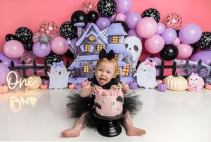 Baby girl in black tutu celebrates first Halloween birthday smash cake with purple ghosts and balloon backdrop.