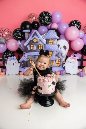 Baby girl in black witch tutu smashes Halloween ghost cake surrounded by purple balloons and haunted house decorations.