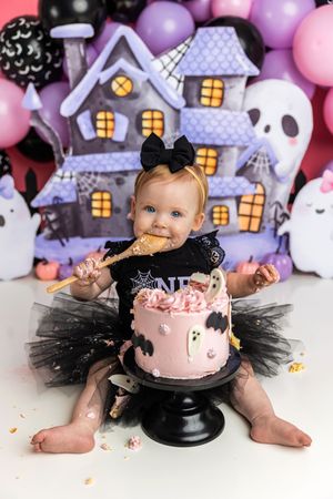 Baby girl in black tutu smashes pink Halloween cake with ghost and bat decorations in front of haunted house backdrop.