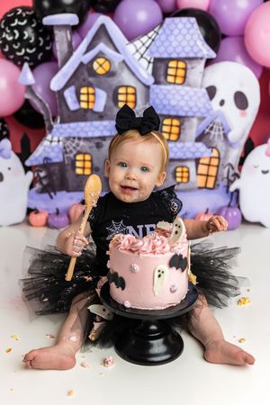 Baby girl in black witch tutu celebrates first Halloween birthday with pink ghost cake and spooky haunted house backdrop.