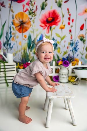 Smiling baby girl in pink top and denim skirt stands at white stool before colorful floral backdrop.