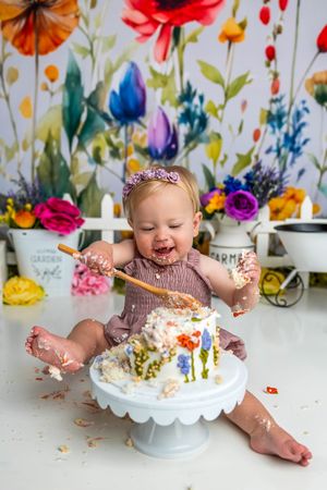 Baby girl smashing her first birthday cake with a spoon, surrounded by colorful floral backdrop and decorations.