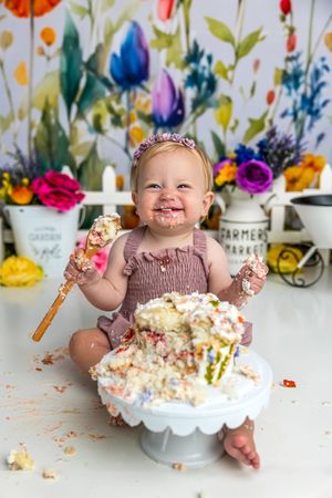 Baby girl smashing her first birthday cake, covered in frosting, holding wooden spoons, sitting in front of colorful floral backdrop.