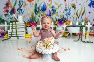 Baby girl smashing her first birthday cake surrounded by colorful floral decorations and white picket fence backdrop.