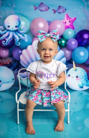 Baby girl in mermaid outfit sits on white chair at underwater-themed first birthday party with colorful balloons.