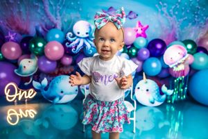 Baby girl in mermaid outfit celebrates first birthday with colorful ocean-themed balloon backdrop and neon One signs.
