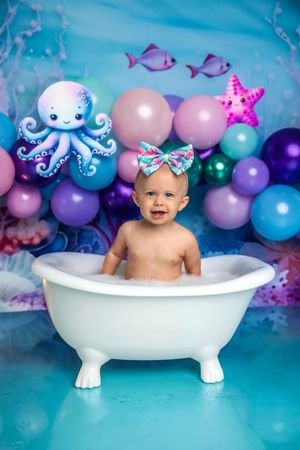 Baby girl in bubble bath with ocean-themed balloons, octopus, starfish decor for first birthday cake smash photo.