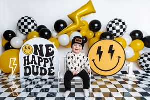 Toddler boy sits on chair at 1st birthday party with smiley face balloons and One Happy Dude sign.