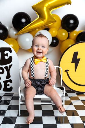 Smiling baby in checkered suspenders and yellow bow tie sits on chair surrounded by black, white, and gold birthday balloons.