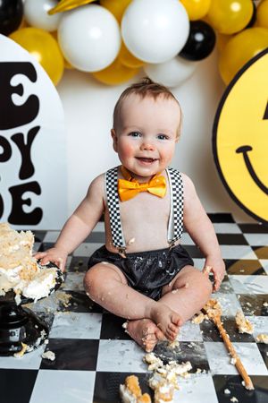 Smiling baby in suspenders and bow tie enjoys a cake smash on a checkered floor with smiley face balloons.