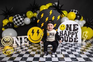 Baby boy sits in front of a smiley face balloon backdrop with ONE HAPPY DUDE signs for his first birthday photo.