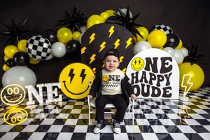 Baby boy sits among yellow balloons and smiley face decorations at a One Happy Dude first birthday party.